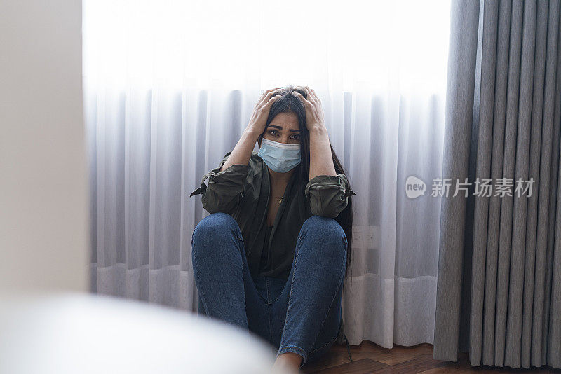 一名妇女坐在房子的地板上，态度非常无聊，因为她是COVID - 19阳性，必须用口罩隔离，以避免病毒的传播。